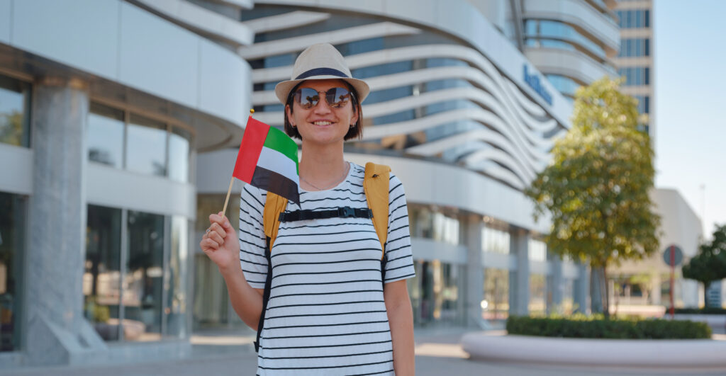 Happy young asian female traveler with backpack and hat with UAE flag against scenic skyscrapers in Abu Dhabi. Immigration, student education and arab emirates citizenship concept